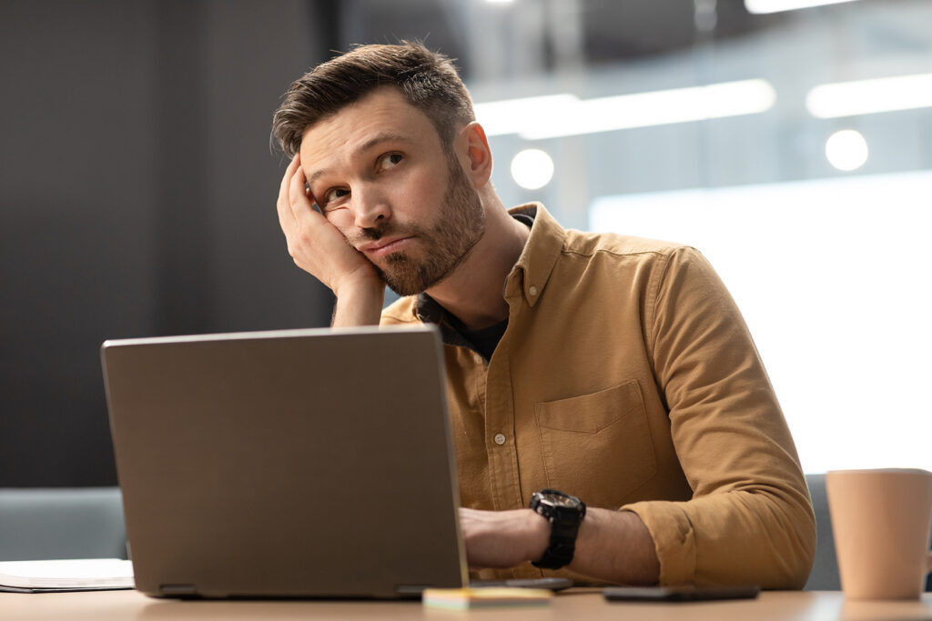 A man is shown sitting in front of a computer, head leaning against one of his hands. He looks unhappy.