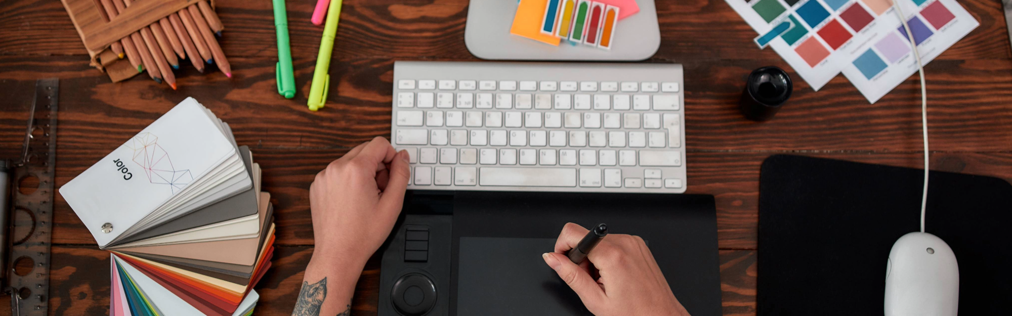 A top-down view of a graphic designer's desk. You see hands poised over a drawing tablet, a keyboard, a computer mouse, and color swatches.
